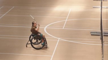 Tekerlekli sandalyedeki beyaz bir kızın, kapalı sahada görünmez bir atletle badminton oynarken geniş açılı çekimi.