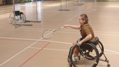 Yüksek açılı, sarı saçlı, beyaz tenli, tekerlekli sandalyede badminton oynayan, kapalı sahada raketle servis yapan bir sporcu.