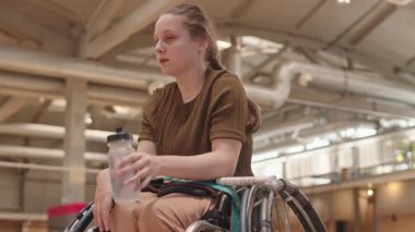 Sarışın, beyaz, tekerlekli sandalyede elinde badminton raketi, boynunu uzatmış, kapalı alanda su içen genç bir kız.