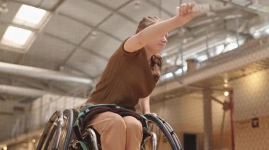 Sarışın, beyaz tenli, tekerlekli sandalyede badminton oynayan, kapalı sahada raketle servis yapan...