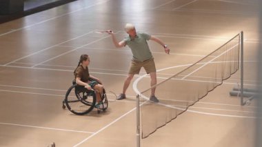 Yüksek açılı beyaz saçlı erkek beyaz koçun badminton raketiyle iç sahadaki tekerlekli sandalyedeki genç bayana yaptığı hareketleri gösteren uzun açılı bir görüntü.
