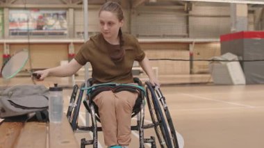 Kafkasyalı engelli bir kızın jimnastik salonunda sırtı spor tekerlekli sandalyeye dönük ısınması, sonra da badminton raketiyle gitmesi.