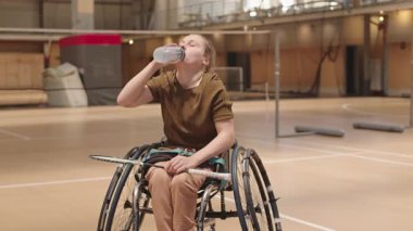 Orta boylu, sarışın, tekerlekli sandalyede elinde badminton raketi olan, kapalı alanda su içen beyaz kız.