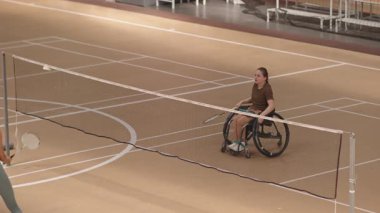 Tekerlekli sandalyedeki beyaz bir kızın, kapalı sahada tanınmayan bir sporcuyla badminton oynarken geniş açılı görüntüsü.
