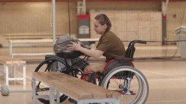 Beyazlı engelli bir kızın spor salonunda bankta durup sırt çantası takıp spor tekerlekli sandalyeye binerken fotoğrafını çekmek.