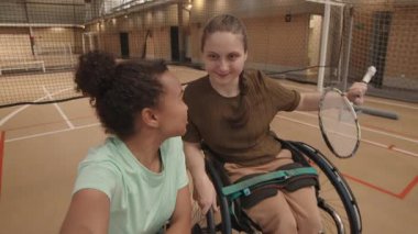 Elinde badminton raketi tutan, konuşan, gülümseyen ve iç sahada kameraya bakan on beş yaşında, beyaz tekerlekli sandalyede oturan genç ve çok kültürlü bir bayan sporcunun el bilgisayarı.