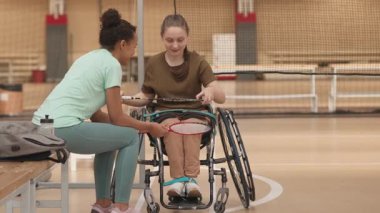 Spor tekerlekli sandalyedeki beyaz bir kızın spor salonunda kadın partnerle konuşması, sonra ikisinin de badminton raketi ile gitmesi.