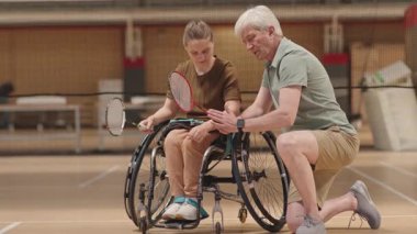 Son sınıf Kafkas koçu tekerlekli sandalyedeki genç bir kızın yanında diz çöküp ona içeride raketle badminton öğretiyor.