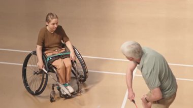 Beyaz saçlı koçun, badminton raketiyle iç sahada tekerlekli sandalyedeki beyaz kadın ergene yaptığı yüksek açılı çekim.