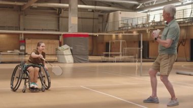Geniş açılı Kafkas koçu, kapalı sahada duruyor, tekerlekli sandalyedeki genç kıza mekikleri fırlatıyor, badminton raketiyle servis yapıyor.