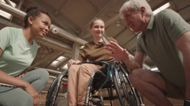 Beyaz saçlı beyaz adam, çok ırklı genç sporcu kadın ve tekerlekli sandalyedeki genç kız kapalı mekanda el ele tutuşuyorlar.