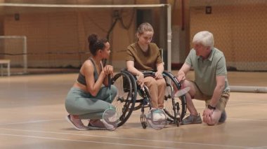 Beyaz saçlı beyaz saçlı beyaz erkek koç ve çok ırklı bayan sporcunun tekerlekli sandalyede genç bir kızın yanında diz çöküp kapalı alanda badminton maçı stratejisini tartışmaları.