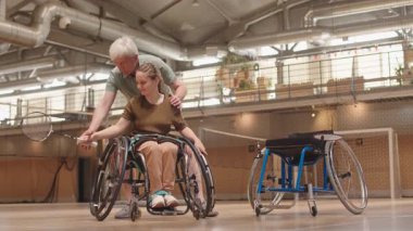 Kafkas koçu, engelli genç bir kızın tekerlekli sandalyesinin arkasında duruyor. Salonda raketle ona badminton el hareketleri öğretiyor.