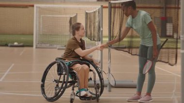 Tekerlekli sandalyedeki on beş yaşındaki beyaz bir kızın kapalı sahada çok ırklı kadın rakiplerle el sıkışması, badminton raketleri tutan sporcular.
