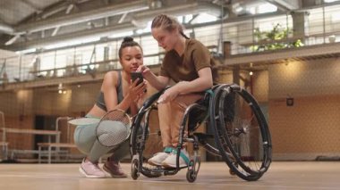 Düşük açı, tekerlekli sandalyede oturan, çok ırklı bayan sporcuyla konuşan, spor salonunda badminton raketleri tutan arkadaşlarıyla konuşan,