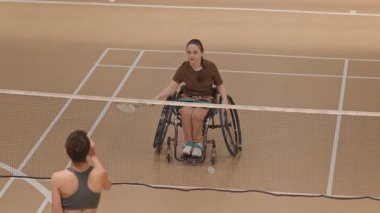 Yüksek açılı tanınmayan bir sporcu, ağdan top fırlatıyor, tekerlekli sandalyedeki beyaz kız servis atıyor, iç sahada gülümsüyor.