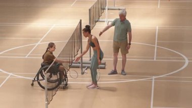 Yüksek açı, genç çok ırklı kadının badminton maçından sonra tekerlekli sandalyedeki beyaz kızla tokalaşırken görüntüsü.