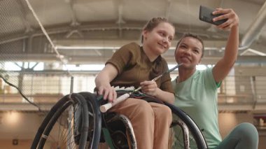 Düşük açılı, orta boylu, mutlu, çok ırklı bir kadının, tekerlekli sandalyede, elinde badminton raketi tutan genç beyaz bir kızla akıllı telefonuyla selfie çektiği bir film.