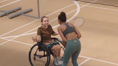 Tekerlekli sandalyedeki çok kültürlü genç kadın ve okul çağındaki kızın kapalı sahadaki maçı kazandıktan sonra beşlik çaktığı ve konuştuğu yüksek açılı bir çekim.