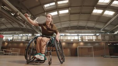 15 yaşında, tekerlekli sandalyede badminton oynayan, kapalı sahada raketle mekiğe çarpan beyaz bir kız.