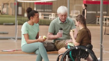 Orta boylu, yaşlı, beyaz bir erkek ve çok ırklı bir kadının spor salonundaki kadın sporcuyla konuştuğu ve meyve yediği bir spor salonu.