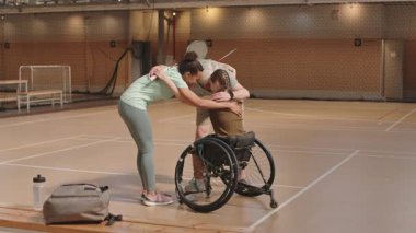 Son sınıf beyaz adam, genç çok ırklı sporcu kadın ve tekerlekli sandalyedeki genç kızın maçtan önce spor salonunda kucaklaşması.