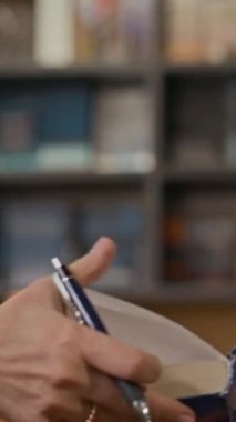 Orta yaşlı beyaz kadın yazarın dikey gülümsemesi yerel kitapçıda edebiyat dersinde kalemle kitap imzalarken.