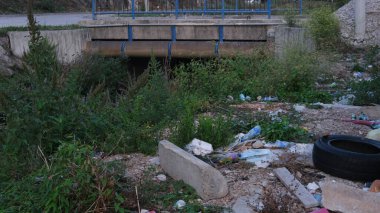 Beton köprünün yanındaki yol kenarı çöplük, plastik şişeler ve eski bir lastik dolu.. 