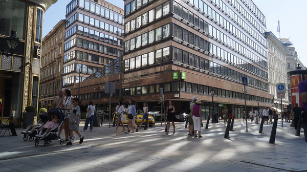 People walk past modern and historic buildings on a busy city street on a sunny day