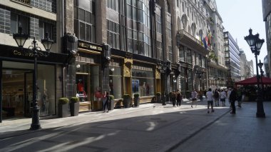 Yayalar güneşli bir caddede logoları olan mağazaların önünden geçiyorlar.