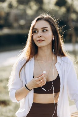 Beautiful girl with freckles in white shirt sits in park on bench and listens to music in headphones