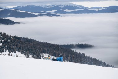 Snowboarder in jacket stands on top of mountain and posing to camera