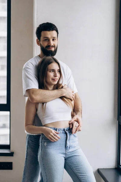 Young cute couple in white t-shirts and blue jeans stand in new apartment and rejoice in happiness