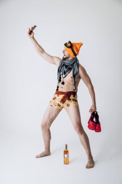 Şapkalı ve kayak gözlüklü bir adam ellerinde sarı lastik eldivenler ve pembe kadın ayakkabıları ile selfie çekiyor. 