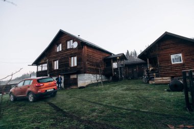Turuncu SUV, dağlar ve ahşap bir evin arkasında duruyor.