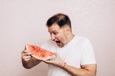 Beyaz tişörtlü adam olgun karpuz tutuyor ve yiyor. Bahçeden yaz meyveleri