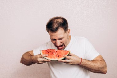 Beyaz tişörtlü adam olgun karpuz tutuyor ve yiyor. Bahçeden yaz meyveleri