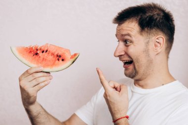 Beyaz tişörtlü adam olgun karpuz tutuyor ve yiyor. Bahçeden yaz meyveleri