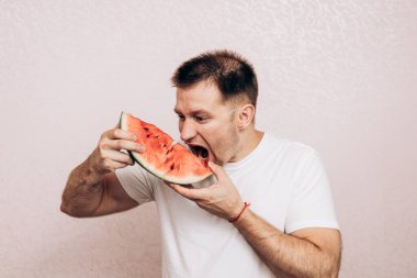 Beyaz tişörtlü adam olgun karpuz tutuyor ve yiyor. Bahçeden yaz meyveleri