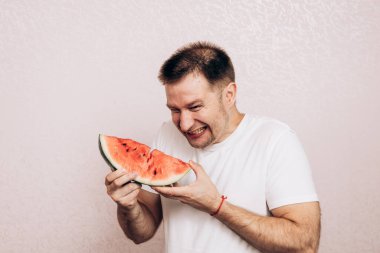 Beyaz tişörtlü adam olgun karpuz tutuyor ve yiyor. Bahçeden yaz meyveleri