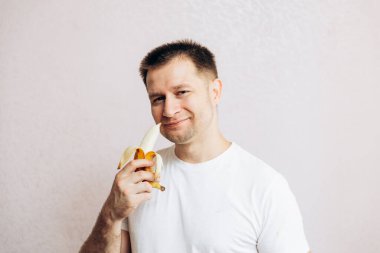 Beyaz tişörtlü adam olgun sarı muzu tutuyor ve yiyor. Bahçeden yaz vitamini meyveleri