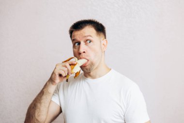 Beyaz tişörtlü adam olgun sarı muzu tutuyor ve yiyor. Bahçeden yaz vitamini meyveleri