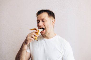 Beyaz tişörtlü adam olgun sarı muzu tutuyor ve yiyor. Bahçeden yaz vitamini meyveleri