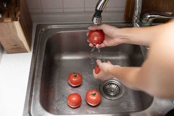 Women's hands wash red tomatoes in sink. Peeling tomatoes under water in kitchen
