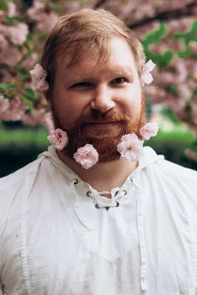 Portrait of man with red beard with pink flowers in it. Gender equality. Spring came