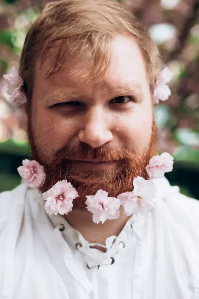 Portrait of man with red beard with pink flowers in it. Gender equality. Spring came