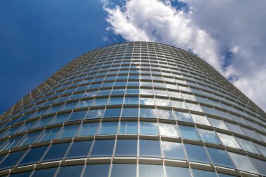 Kavisli cam gökdelen mavi gökyüzünü ve beyaz bulutları yansıtıyor..