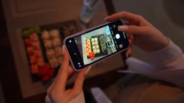 Kız, rahat Japon restoranında akıllı telefon kullanarak suşi fotoğrafı çekiyor. Kadın eli, modern akıllı telefonla yiyeceklerin fotoğrafını çeker. Yaklaş, 4K