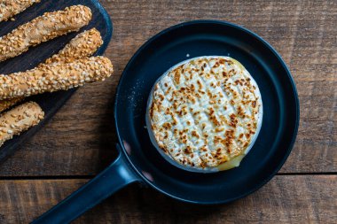 View of baked on frying pan camembert cheese and bread sticks with sesame seeds on wooden table background, close up, top view