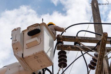 Male electrician standing in mobile crane basket and repairs a wire of the power electrical line on sky background , Thailand